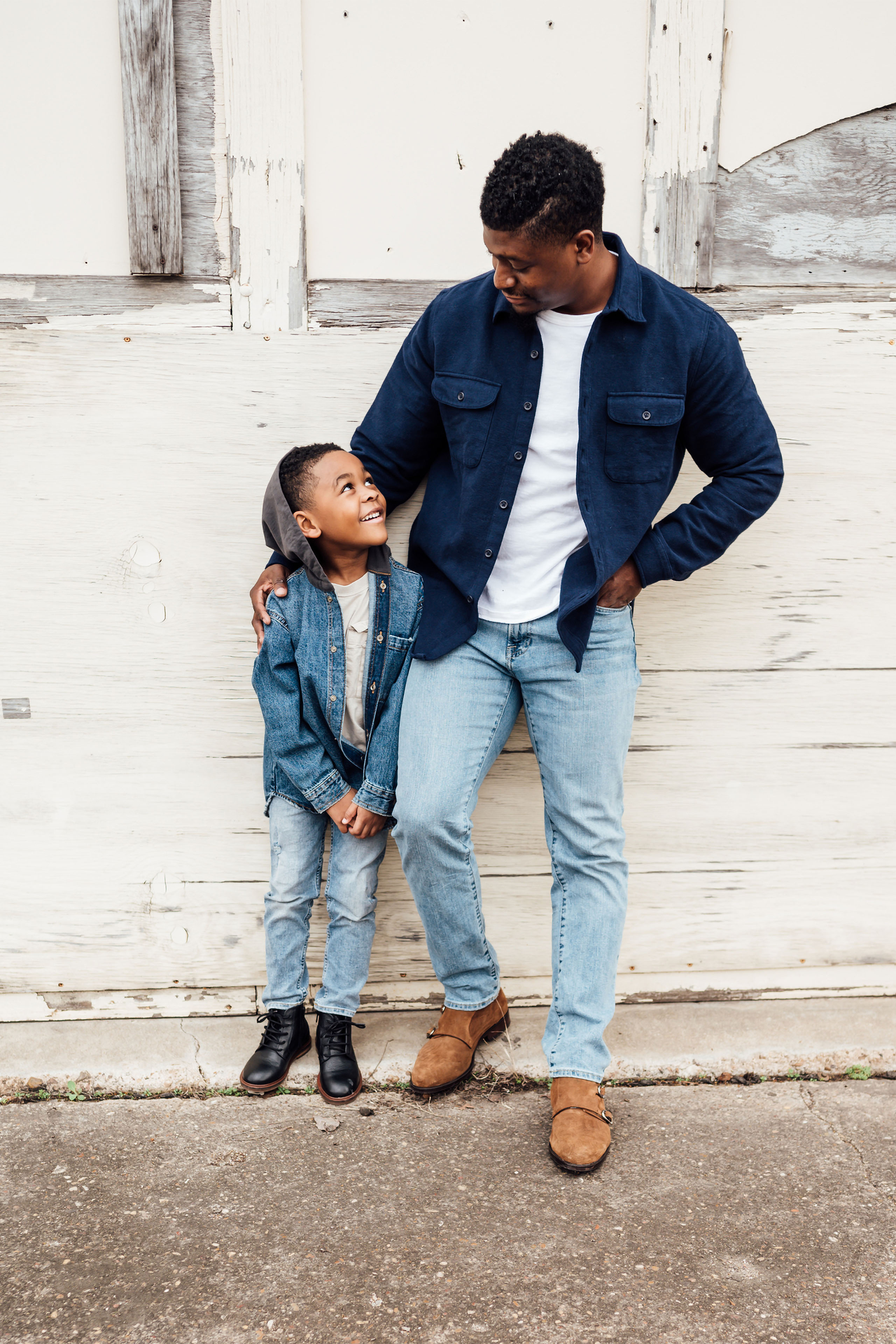 A father and his young son leaning against a white wall, smiling at each other.