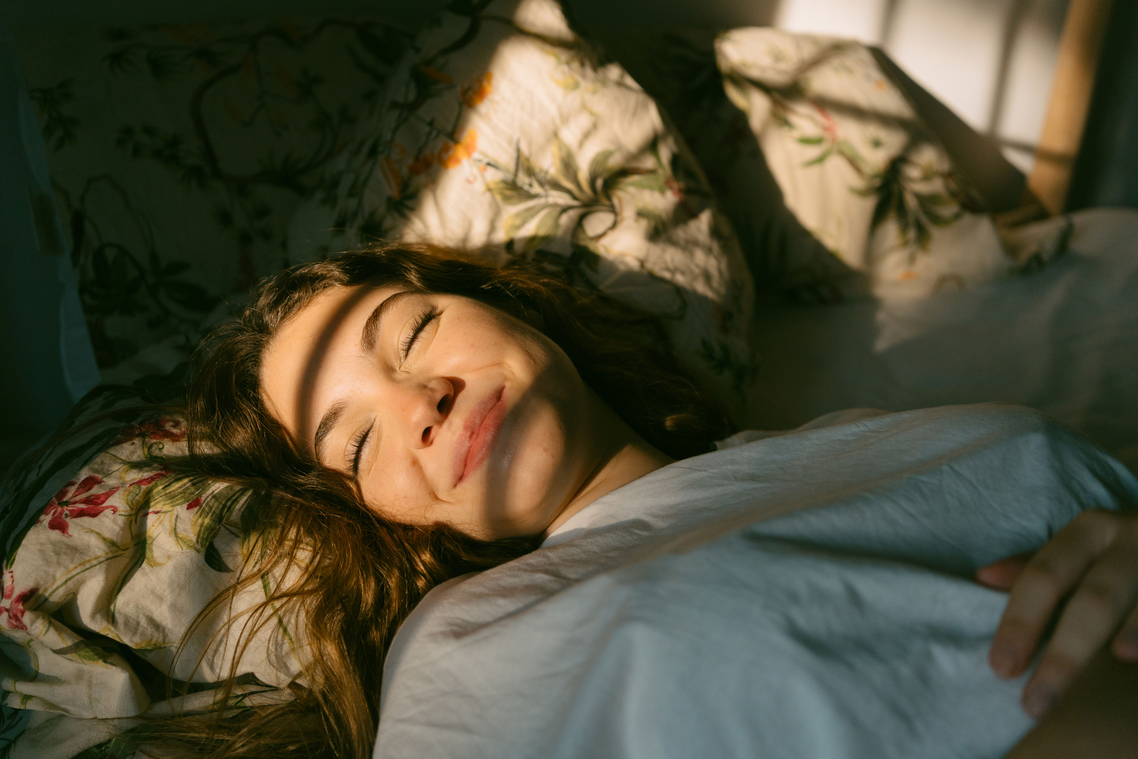 A woman lying in bed in morning light, smiling with her eyes closed.